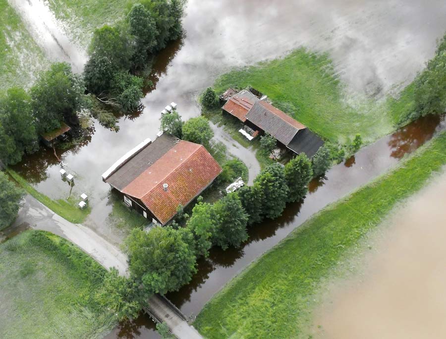 Es ist eine Luftaufnahme zu sehen. Ein Anwesen, auf dem mehrere Gebäude stehen, steht zu großen Teilen unter Wasser. Der Bach, welcher rechts neben dem Haus verläuft und für die Überflutung verantwortlich ist, hat sich bereits wieder in sein Bett zurückgezogen.