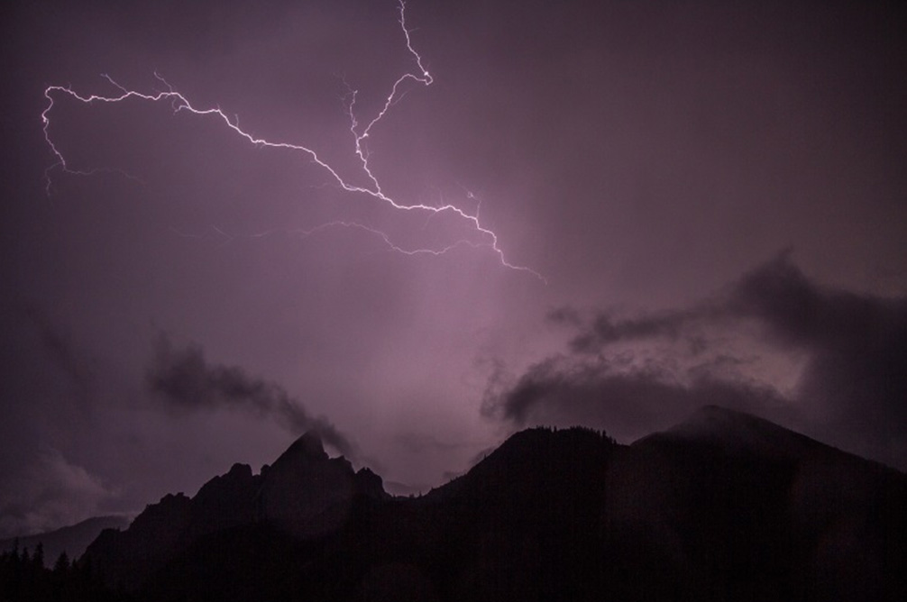 Eine Bergspitze bei Nacht während eines Gewitters. Dabei sind die Gipfel als Silhouette im Vordergrund und im oberen Bildbereich ein Blitz zu sehen.