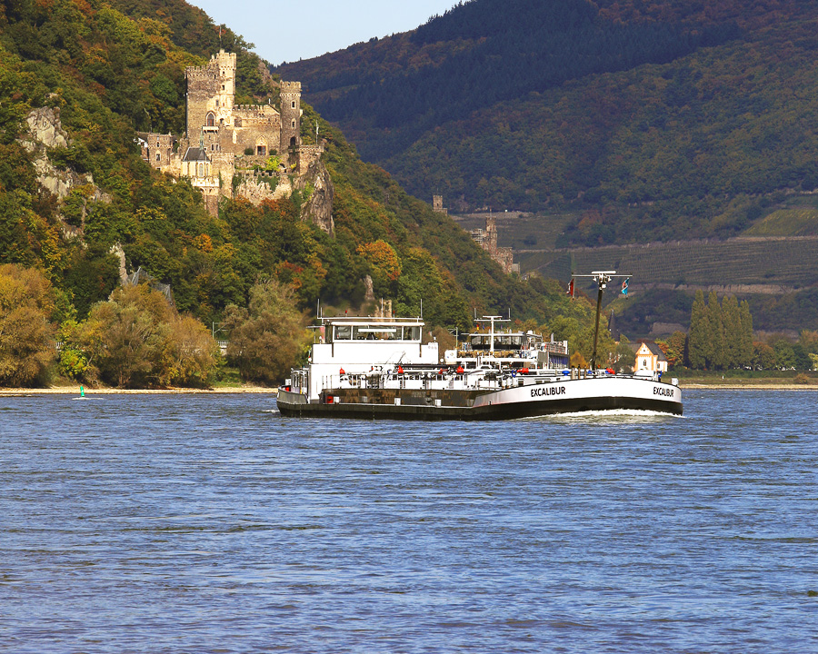 Ein Frachtschiff auf dem Mittelrhein. Im Hintergrund kann man die Talhänge mit einer Burgruine, Wald und Weinbergen erkennen. Im Vordergrund erstreckt sich der Fluss. In der Bildmitte befindet sich ein Frachtschiff auf dem Wasser.