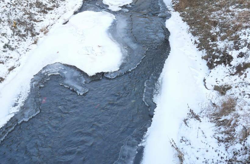 Ein teilweise zugefrorener Bach. Links oben und auf der rechten Seite sind partiell verschneite Uferböschungen zu sehen. In der Bildmitte fließt ein Bach, der am rechten Ufer einen schmalen, schneebedeckten Streifen Eis aufweist und auf der anderen Seite weit in den Bachlauf ragt.