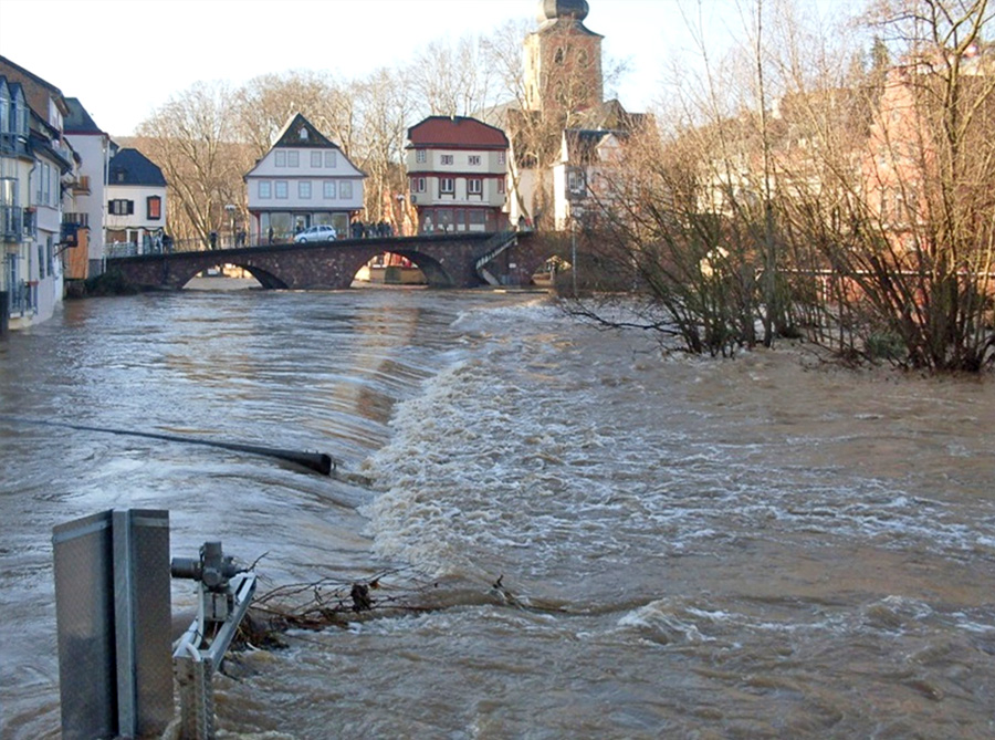 Hochwasser an der Nahe bei Kreuznach. Im Vordergrund fließt die Nahe wild ein Gefälle hinab. Links stehen Häuser im Wasser, rechts ragen Bäume aus dem Wasser. Im Hintergrund sind Brückenhäuser zu sehen.