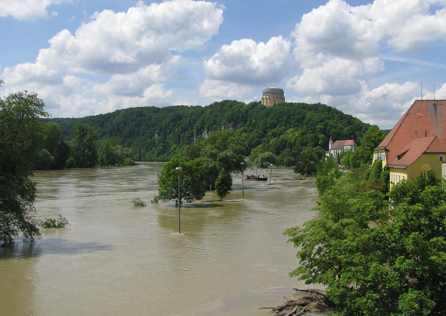 Die Donau, bei Kelheim, bei Hochwasser. Die Donau ist deutlich über die Ufer getreten und hat auch die Gebäude im Bild erreicht. Bäume und Laternenmasten ragen mitten aus dem Wasser. Im Hintergrund ist auf einem Hügel die Befreiungshalle zu sehen.