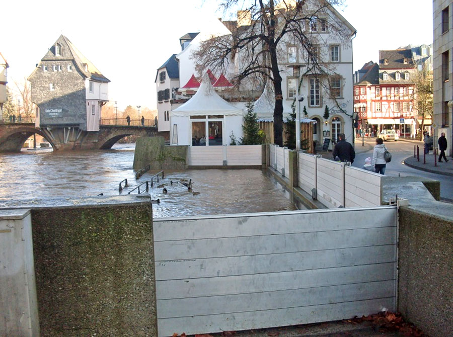 Zu sehen sind Hochwasserschutzmaßnahmen mit mobilen Elementen in Bad Kreuznach an der Nahe. Rechts sind Fußgänger:innen zu sehen, die ohne die Hochwasserschutzmaßnahmen im Wasser stehen würden. Im Hintergrund sind die Brückenhäuser der Alten Nahebrücke zu sehen.