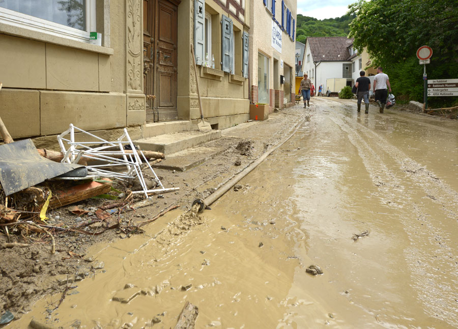 Eine Straße in einer Siedlung nach einer Sturzflut. Auf der linken Seite befindet sich ein Häuserzug und rechts oben im Hintergrund sind Passanten sichtbar. Im Vordergrund ist eine von Schlamm bedeckte Fahrbahn und angeschwemmtes Treibgut zu sehen.