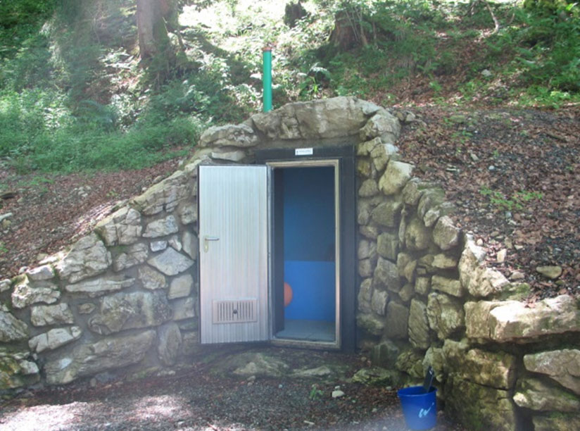 Außenansicht einer Quellmessstelle. Die Messstelle befindet sich in einem kleinen Raum, der in einen Hang im Wald mit Steinen eingelassenen ist. Die Tür zum Raum mit der Messstelle ist geöffnet. Von außen ist ein blaues Becken zu erkennen.