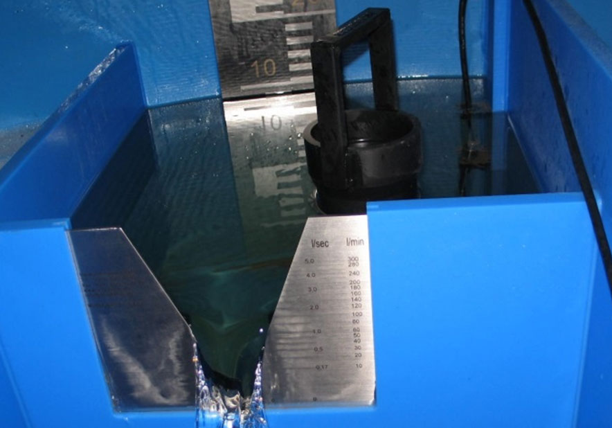 Innenansicht einer Quellmessstelle: Messtechnik. Diese besteht aus einem oben offenen, quadratischen Plastikbehälter, in den das Wasser aus der Quelle hineinfließt. An der hinteren Gefäßwand befindet sich eine Messlatte, an dem man den Wasserstand im Behälter ablesen kann. In die vordere Seite des Behälters ist eine metallene Messvorrichtung für den Abfluss eingelassen.  An dieser kann man den Abfluss in l/sec und l/min ablesen, während das Wasser wieder aus dem Plastikbehälter fließt.