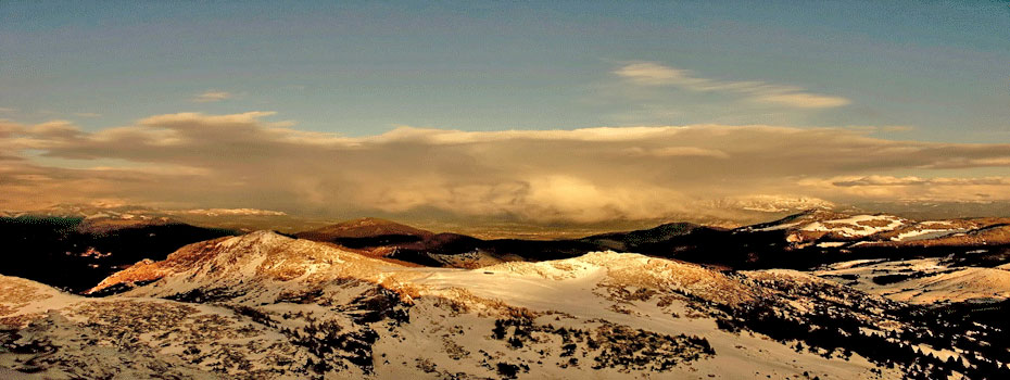 Ein Bergpanorama. Unterhalb des teilweise bewölkten Himmels befinden sich schneebedeckte Berge, die an ihren Gipfeln von der Sonne beschienen werden.